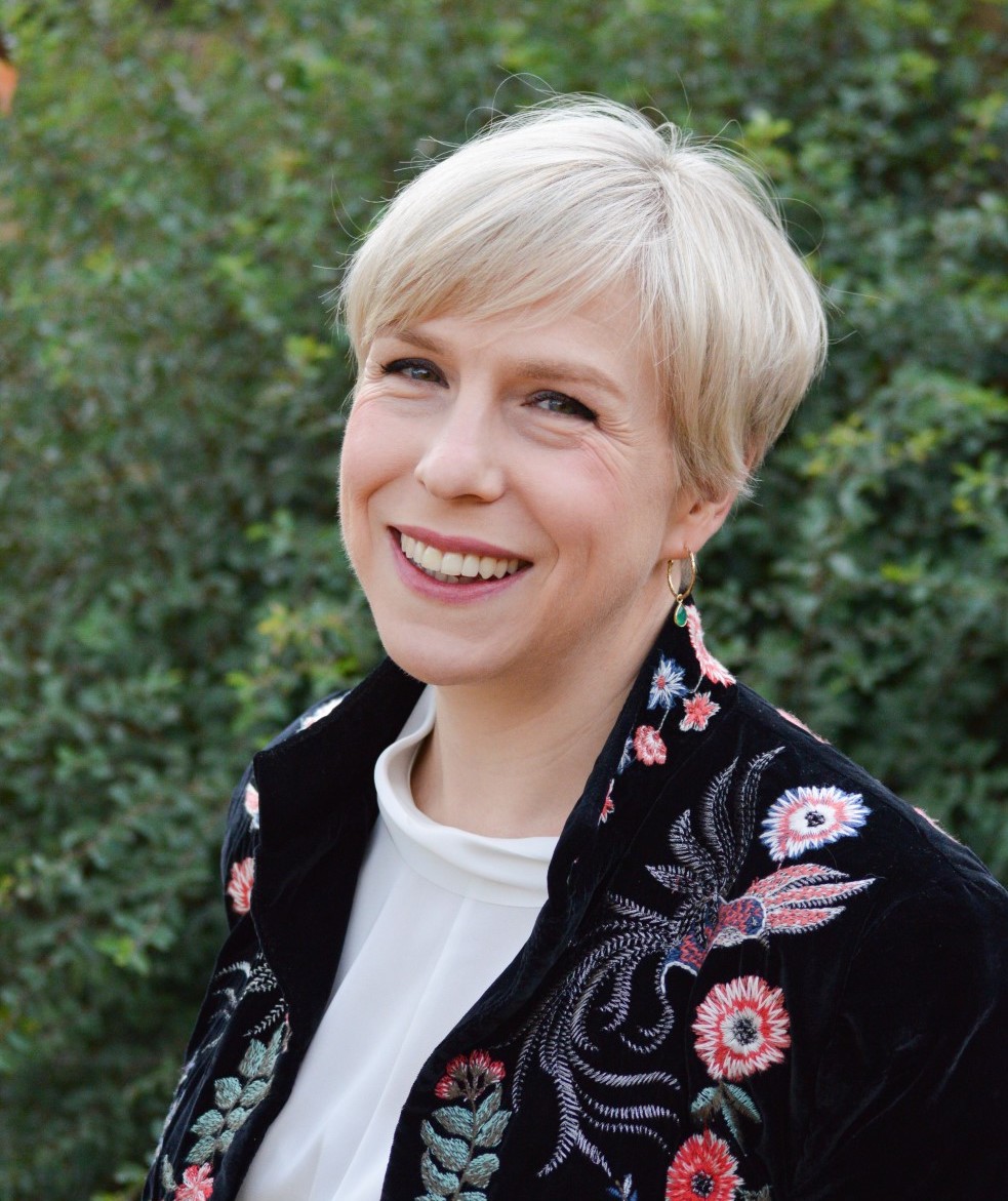 Holly Butcher, CEO of The Unlimited Law, is standing in front of a forest background, wearing a black jacket and smiling.